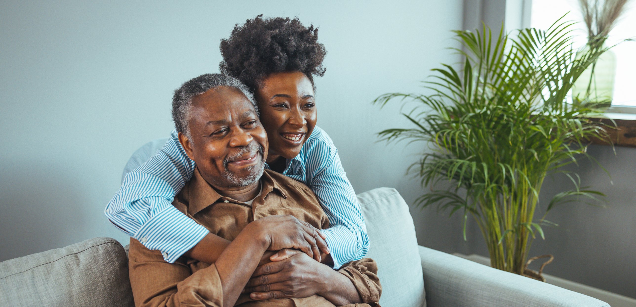 A daughter hugs her father from behind as they sit together at home