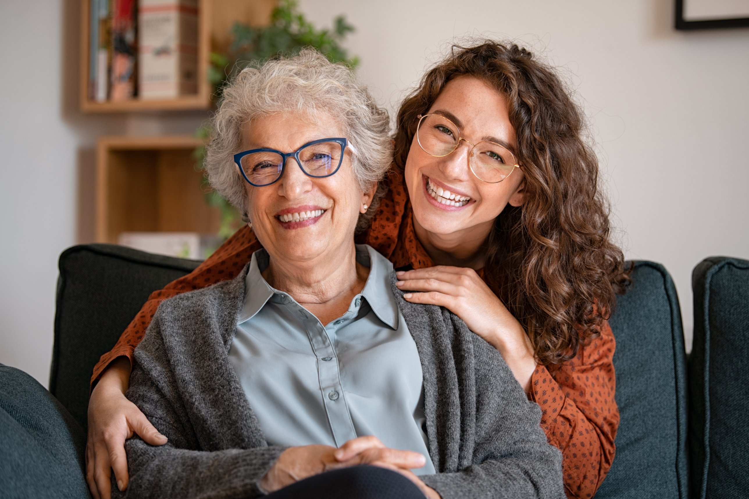 A young woman hugs her smiling grandmother on the living room couch at home